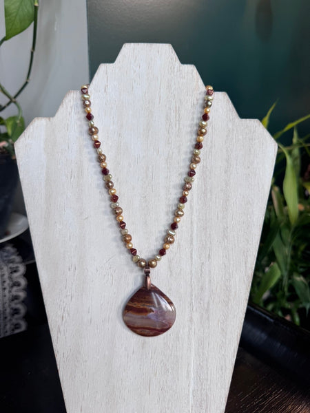 My Two Daughters Jewelry - A beaded necklace with a teardrop-shaped stone pendant displayed on a white jewelry bust. The necklace features small metallic gold beads alternating with dark reddish-brown beads, and the pendant is a polished marbled stone in brown and reddish tones.