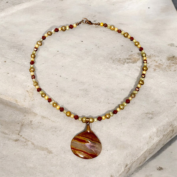 My Two Daughters Jewelry - A beaded necklace with alternating metallic gold and deep red faceted beads, featuring a teardrop-shaped pendant with a marbled brown, gold, and purple pattern, displayed on a light stone surface.