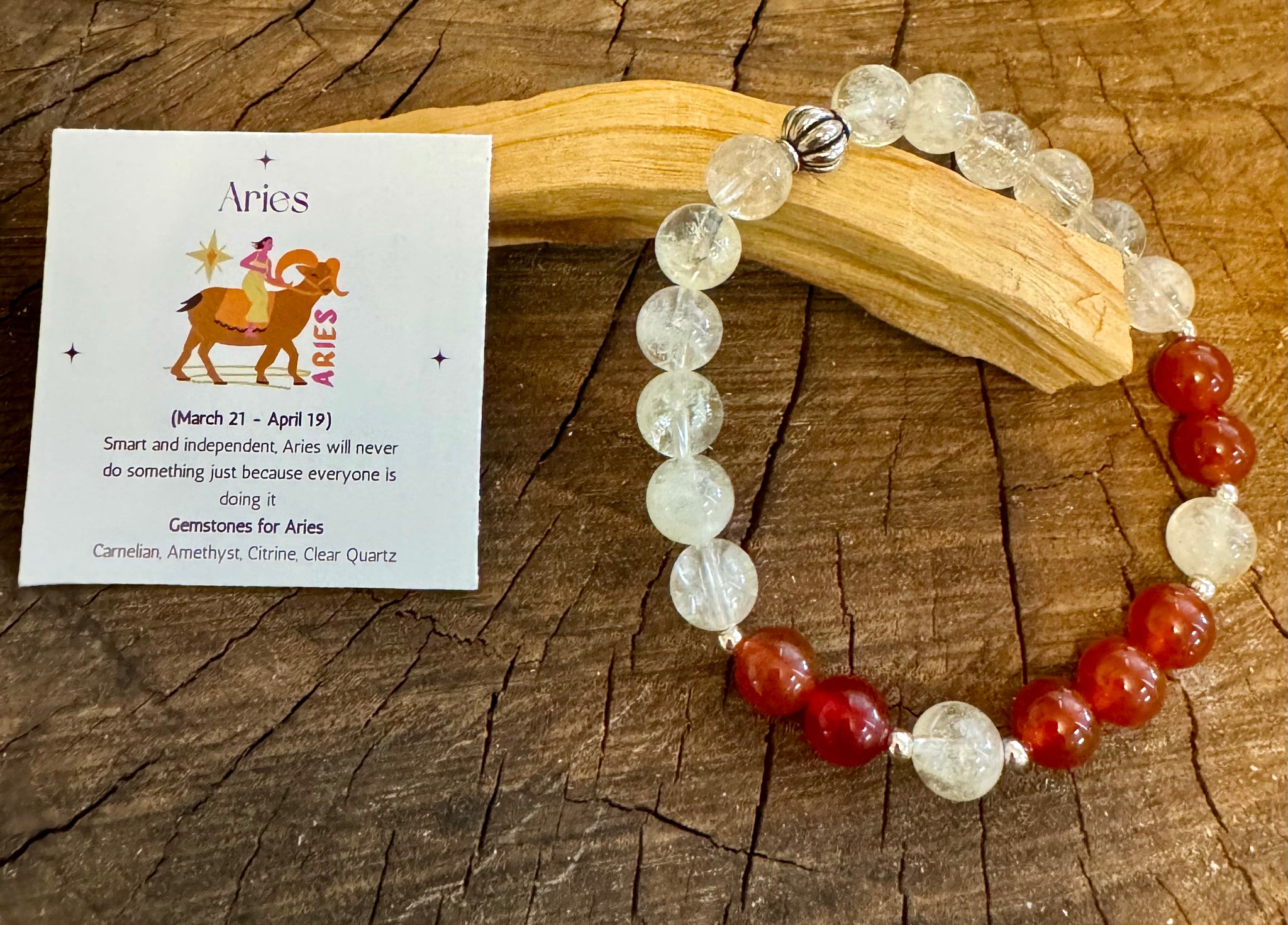 My Two Daughters Jewelry - An Aries bracelet featuring clear quartz and red carnelian beads, displayed on a wooden surface with a zodiac card in the background.