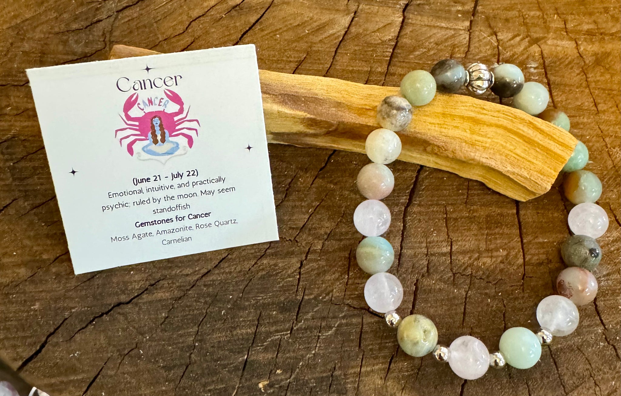 My Two Daughters Jewelry - A bracelet with round beads in shades of green, pink, and brown, along with geometric patterned beads, displayed on a wooden surface next to a card with the Cancer zodiac symbol and a brief description.