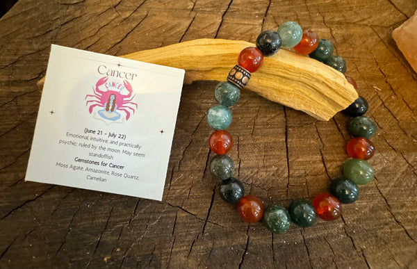 My Two Daughters Jewelry - A bracelet with round beads in shades of green, pink, and brown, along with geometric patterned beads, displayed on a wooden surface next to a card with the Cancer zodiac symbol and a brief description.