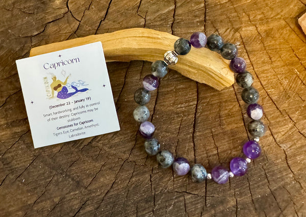 My Two Daughters Jewelry - A colorful gemstone bracelet laid out on a wooden branch, with a card depicting the Capricorn zodiac symbol and a brief description of the product.