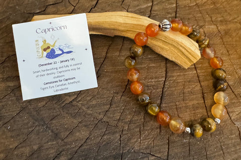 My Two Daughters Jewelry - A colorful gemstone bracelet laid out on a wooden branch, with a card depicting the Capricorn zodiac symbol and a brief description of the product.