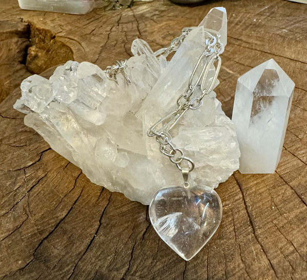 My Two Daughters Jewelry - A clear quartz heart-shaped necklace pendant hanging over a cluster of clear quartz crystals on a wooden surface.