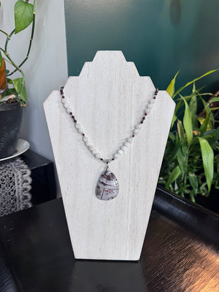 My Two Daughters Jewelry - A beaded necklace with a teardrop-shaped stone pendant displayed on a white jewelry stand. The necklace features white round beads, small dark red beads, and a central marbled gray and white teardrop pendant with red veining, set against a dark table with green plants in the background.