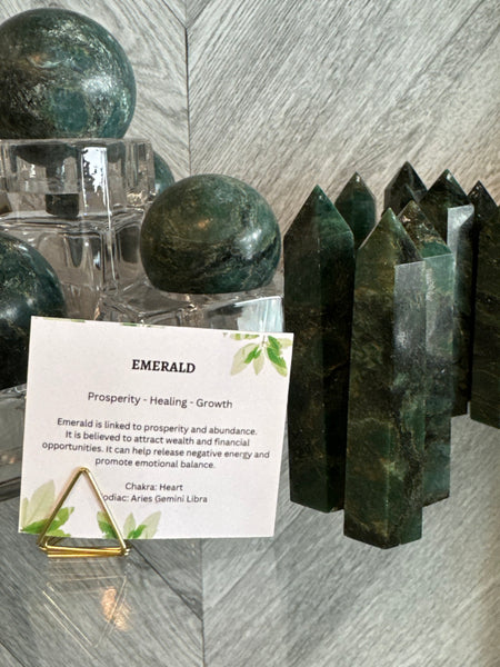 Kelowna Crystal Cave - A display of emerald crystals, including polished green spheres, pointed green crystal towers, and a small white informational card on a gold stand, set against a gray wood-grain background.