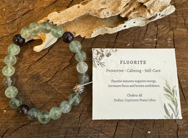 My Two Daughters Jewelry - A green and purple fluorite bracelet displayed on a wooden surface, with a card describing its properties as protective, calming, and self-care related to the chakra and zodiac.