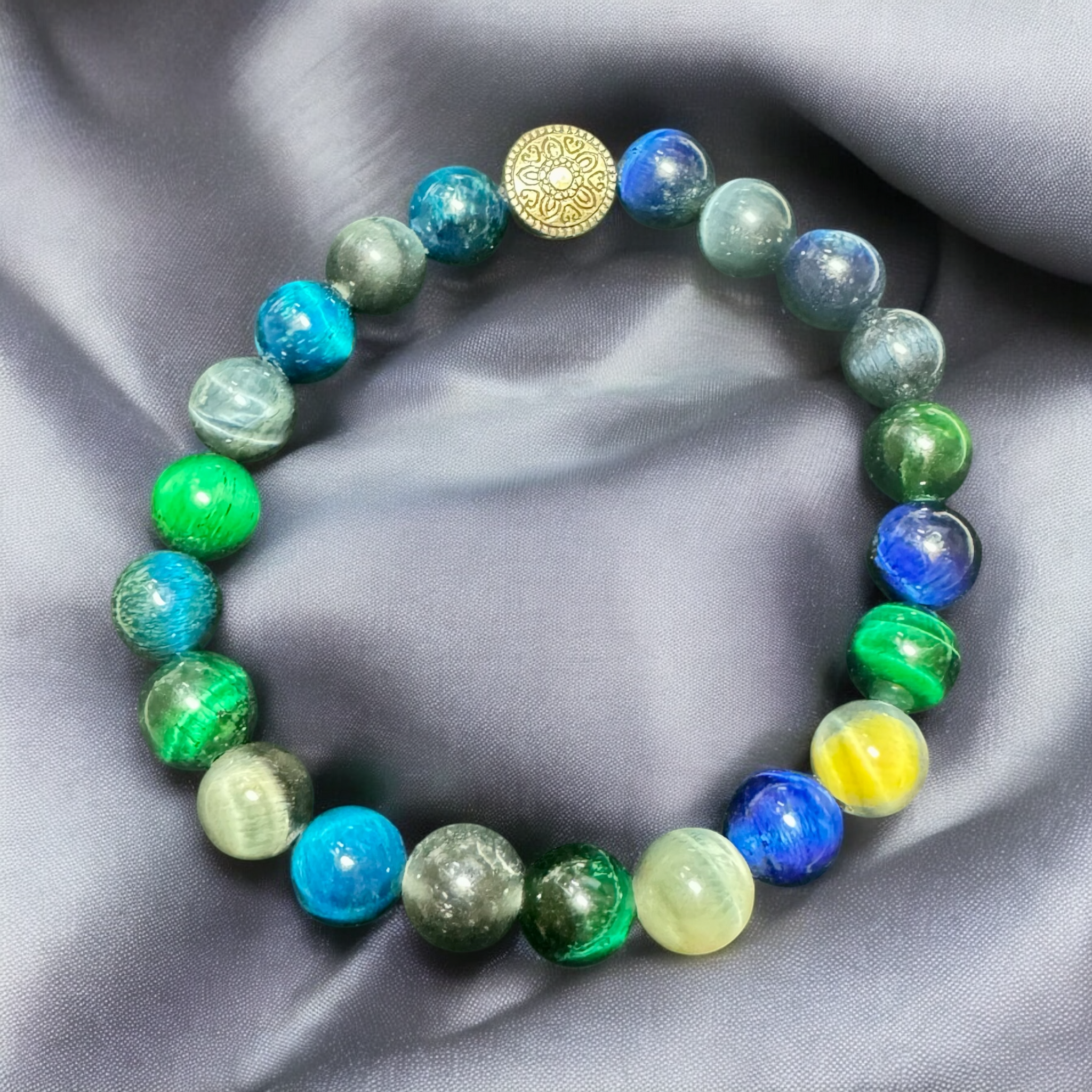 My Two Daughters Jewelry - A beaded bracelet with green and blue colors, featuring a golden spherical charm, displayed on a silky gray background.