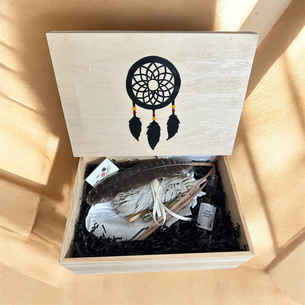 TRUE ART KELOWNA - An open wooden gift box with the lid printed with a black dreamcatcher design, filled with a white sage bundle, a brown feather, and small wrapped items on black shredded paper.