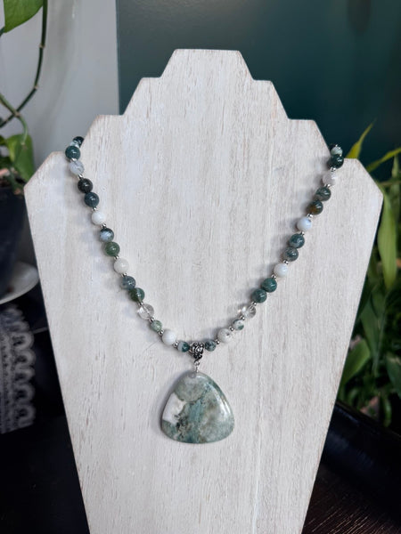 My Two Daughters Jewelry - A beaded necklace with a large teardrop-shaped green stone pendant, displayed on a white wooden stand. The necklace features dark green, white, and gray marbled beads with a small silver-toned spacer, set against a dark teal background with blurred green foliage.