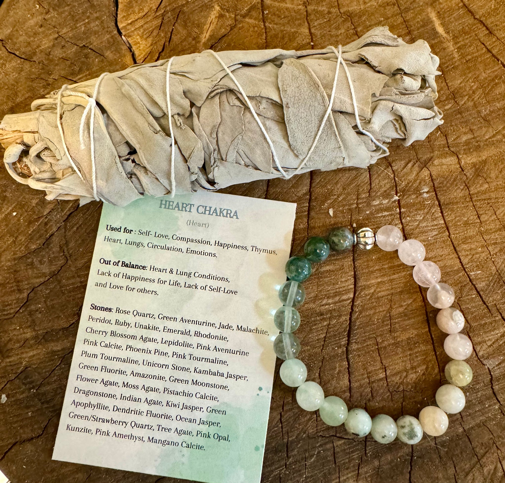 My Two Daughters Jewelry - A variety of semi-precious stone beads strung together, displayed next to a smudge stick and an information card, all laid out on a wooden surface.