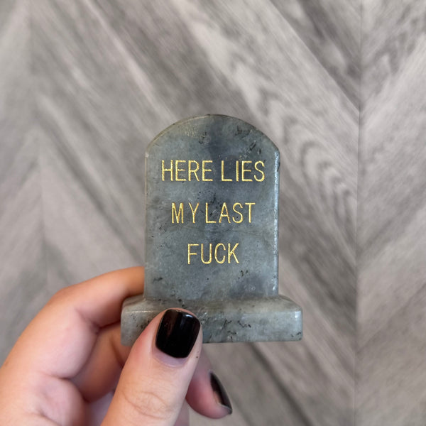 Kelowna Crystal Cave - Small tombstone with 'HERE LIES MY LAST FU*K' text held by a hand against a gray background