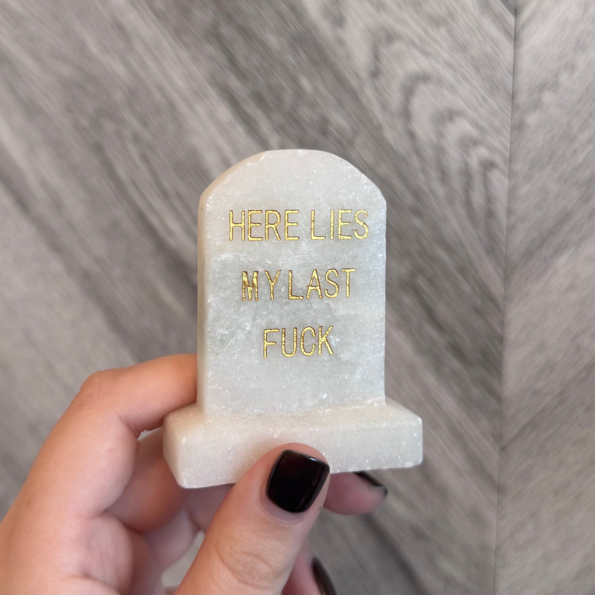 Kelowna Crystal Cave - Small stone tombstone with gold text 'HERE LIES MY LAST FU*K' held by a hand against a wooden background.
