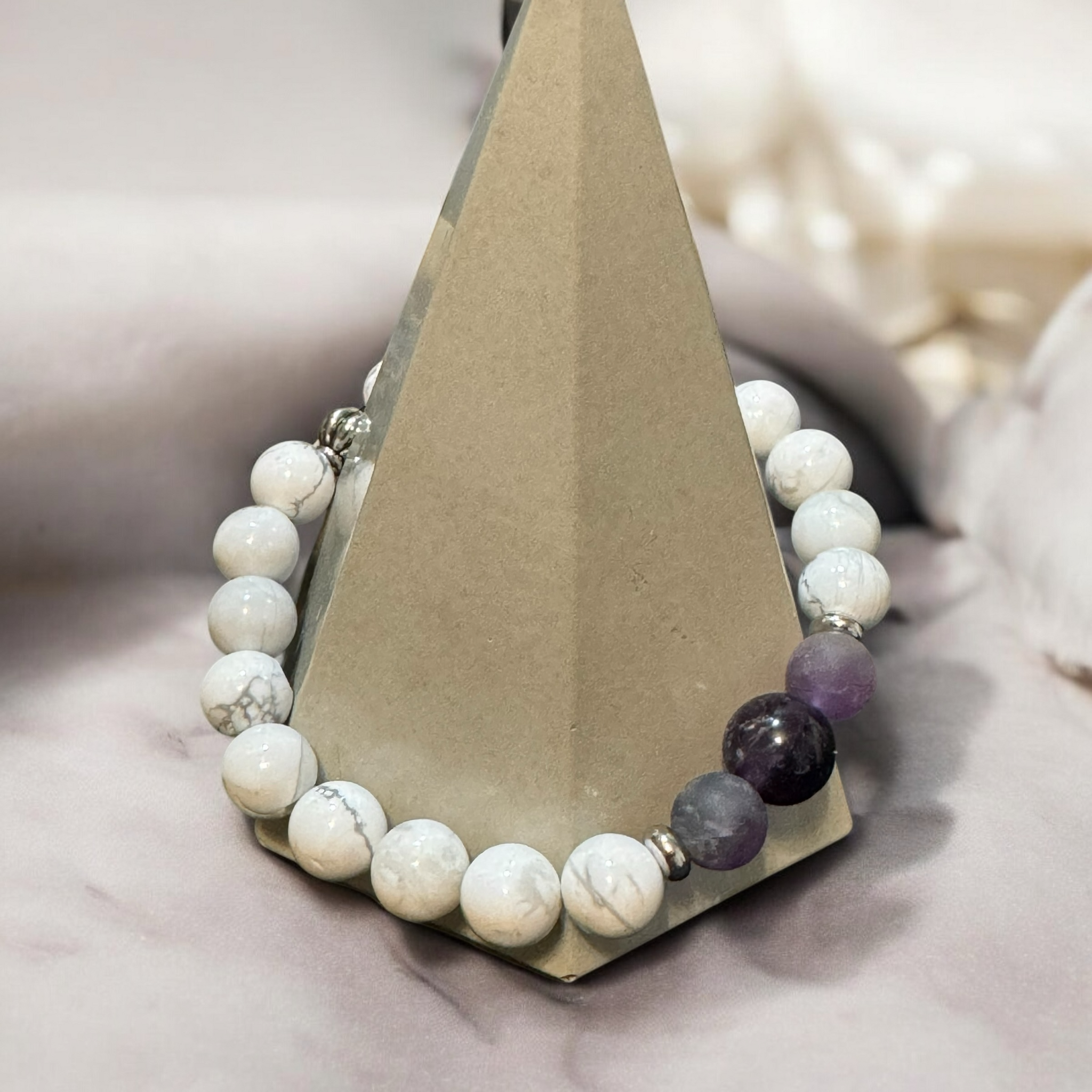 My Two Daughters Jewelry - A bracelet with howlite and amethyst beads displayed on a triangular stand, with a soft, light-colored background.