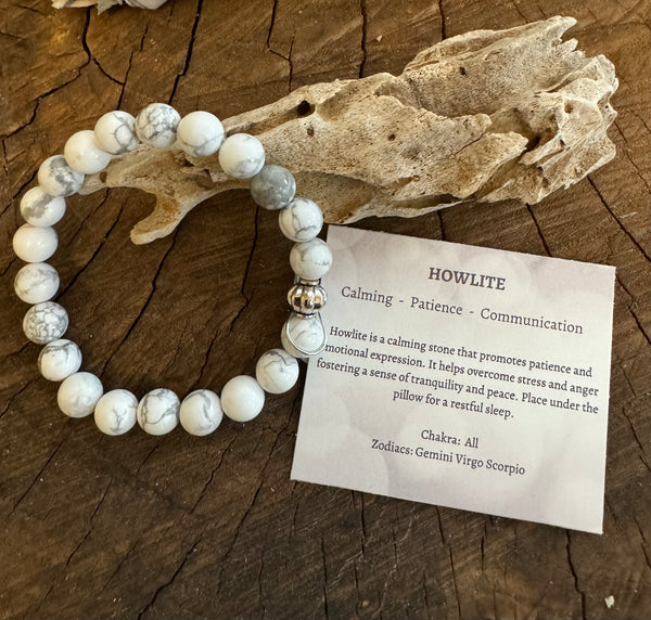 My Two Daughters Jewelry - A white howlite bracelet displayed on a wooden surface, accompanied by a card describing the stone's properties and associations with calming, patience, and communication.