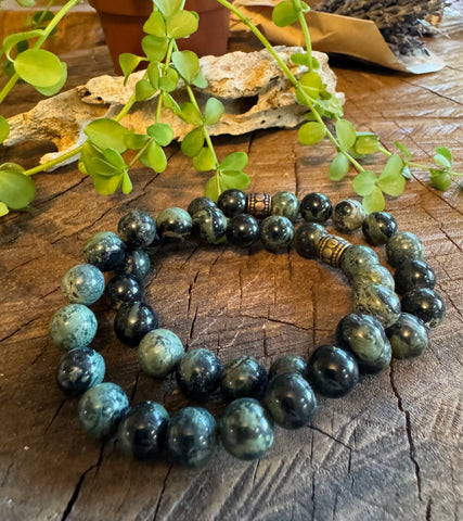 My Two Daughters Jewelry - A Kambaba Jasper Bracelet displayed on a wooden surface with a plant and some decorative items in the background.