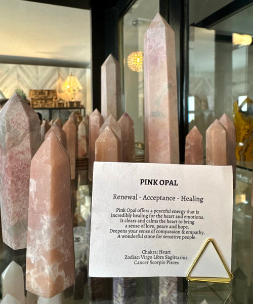 Kelowna Crystal Cave - A pink opal crystal tower displayed on a glass shelf alongside other pink stone points, with a printed card labeled 'PINK OPAL' and a small gold triangular stand in front.