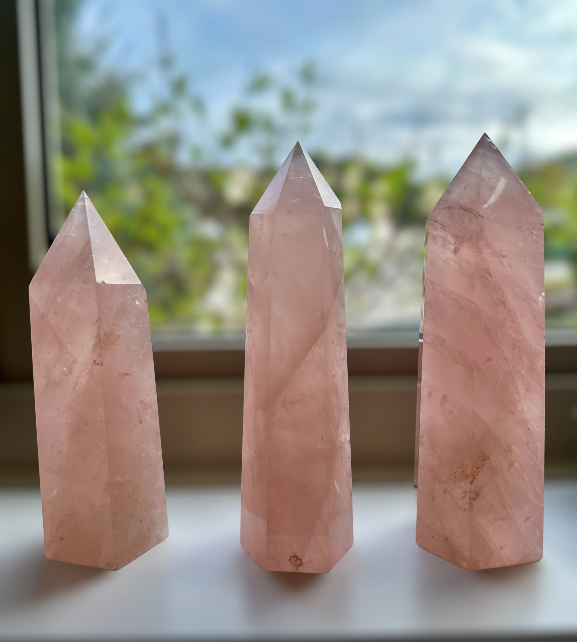 Kelowna Crystal Cave - Three pink rose quartz crystal towers of varying sizes placed on a surface, with a window in the background.