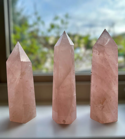 Kelowna Crystal Cave - Three pink rose quartz crystal towers of varying sizes placed on a surface, with a window in the background.