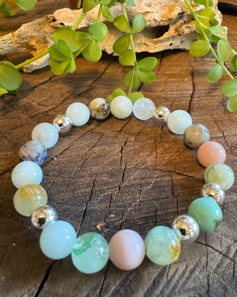 My Two Daughters Jewelry - A bracelet with round beads in shades of green, blue, and pink, with metallic silver accents, displayed on a wooden surface with foliage in the background.