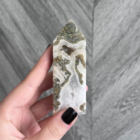 Kelowna Crystal Cave - A hand holding a Moss Agate Tower crystal against a wooden background.