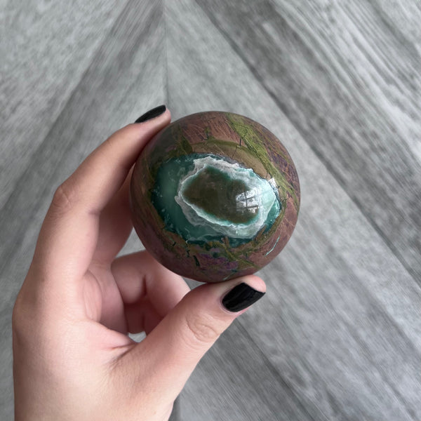 Kelowna Crystal Cave - A hand holding a spherical Nephrite Canadian Jade crystal with a green and brown color pattern.