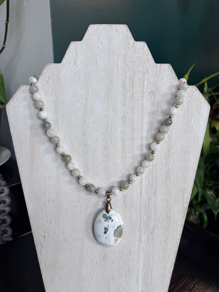 My Two Daughters Jewelry - A beaded necklace with white and light green beads and a white stone pendant, displayed on a white wooden jewelry stand.