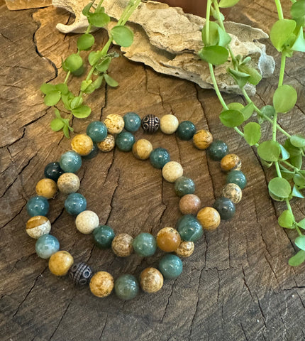 My Two Daughters Jewelry - A bracelet made with beads featuring patterns of green, yellow, and brown, with a decorative metal clasp, displayed on a wooden surface with a piece of green foliage in the background.