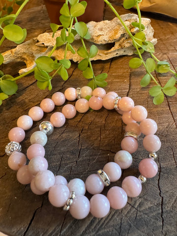 My Two Daughters Jewelry - A pink opal bracelet with silver accents laid out on a wooden surface with green foliage in the background.