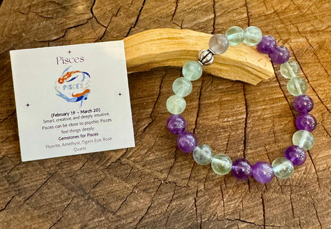 My Two Daughters Jewelry - A colorful bracelet made of fluorite, and Amethyst displayed on a wooden surface, with a card indicating the Pisces zodiac sign and properties of the gemstones.