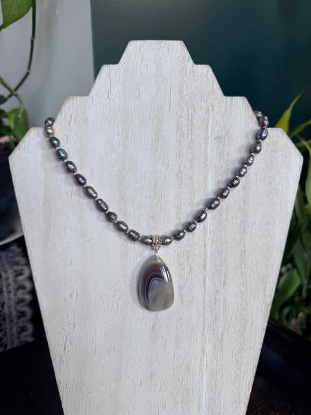 My Two Daughters Jewelry - A beaded necklace with dark gray baroque pearls and a teardrop-shaped agate pendant, displayed on a white wooden jewelry stand.