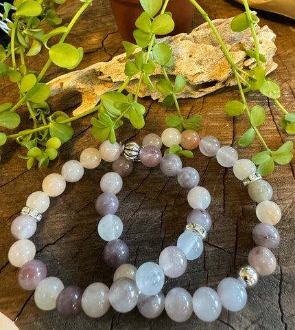 My Two Daughters Jewelry - A bracelet made with purple and translucent beads, featuring metallic accents, displayed on a wooden surface with green leaves in the background.