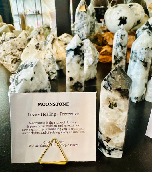 Kelowna Crystal Cave - A Moonstone crystal cluster with a white and black marbled pattern, displayed alongside other crystals and a small gold triangular stand holding a card labeled 'MOONSTONE' with descriptive text.