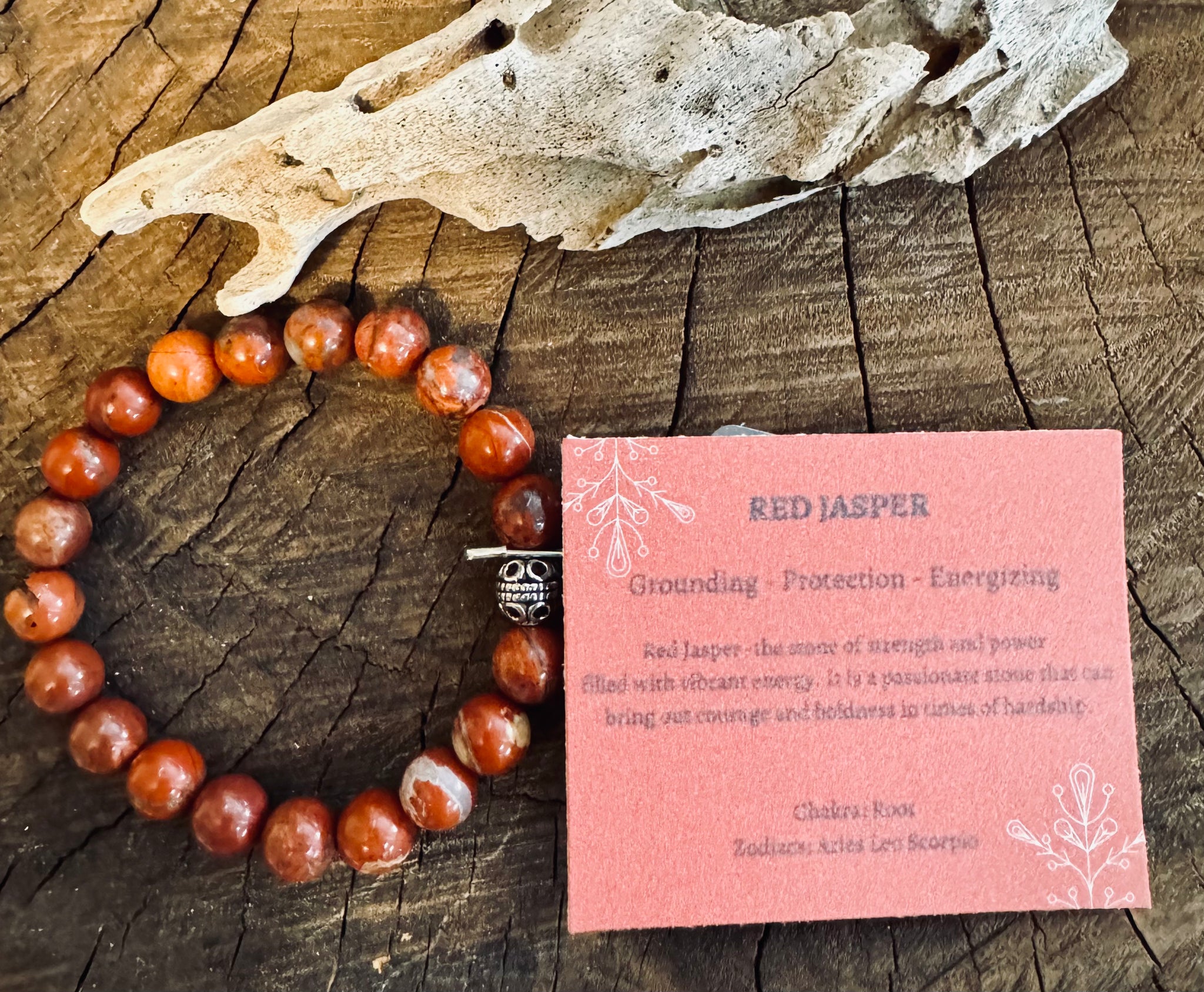 My Two Daughters Jewelry - A red jasper bracelet displayed alongside a wooden piece, with a card detailing the properties of red jasper.