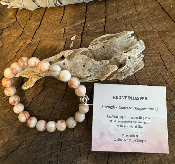 My Two Daughters Jewelry - A Red Vein Jasper bracelet displayed next to a piece of driftwood, with a card describing the stone's properties and meaning.