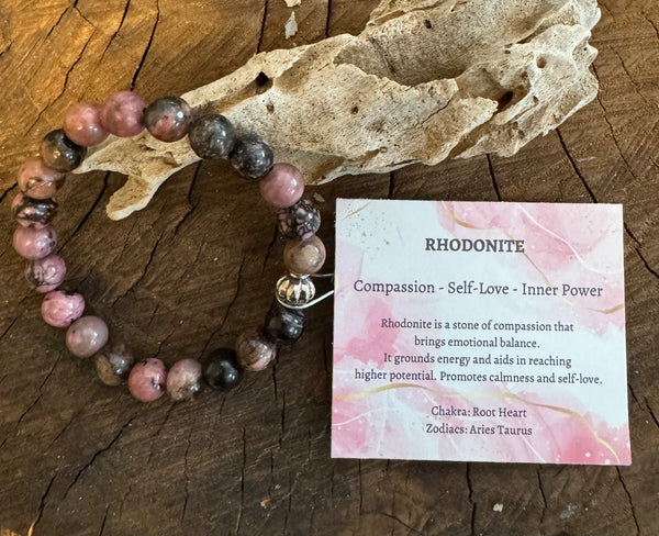 My Two Daughters Jewelry - A Rhodonite bracelet displayed alongside a wooden piece, with a card detailing the properties of Rhodonite, including compassion, self-love, and inner power.