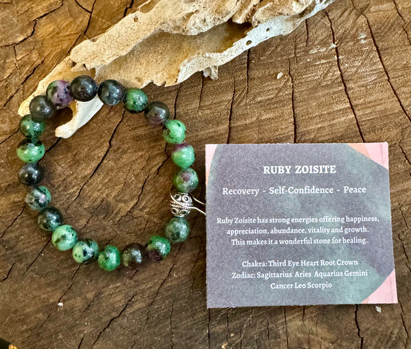 My Two Daughters Jewelry - A Ruby Zoisite bracelet displayed on a wooden surface, with a card describing its properties and meaning.