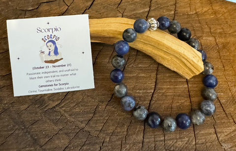 My Two Daughters Jewelry - A Scorpio Bracelet with blue and grey beads displayed on a wooden surface, accompanied by a card with the Scorpio zodiac symbol and a brief description.