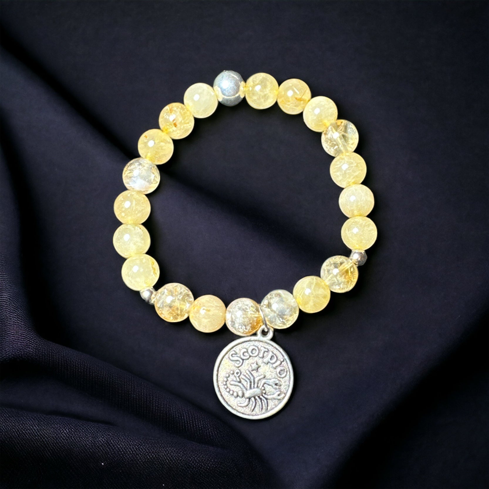 My Two Daughters Jewelry - A bracelet with round yellow beads and a silver-colored Scorpio zodiac sign charm, displayed on a black fabric background.