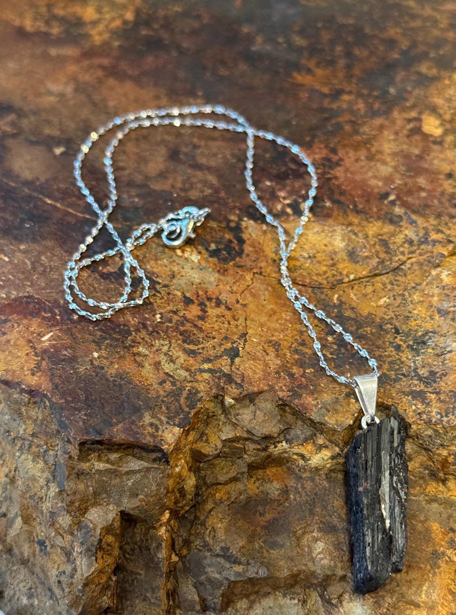 My Two Daughters Jewelry - A black rough tourmaline necklace with a silver chain, displayed against a rock background.