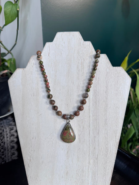 My Two Daughters Jewelry - A beaded necklace with a teardrop-shaped stone pendant displayed on a white jewelry stand. The necklace features small brown and reddish beads with a central silver-toned spacer, set against a dark teal background with blurred green plants.