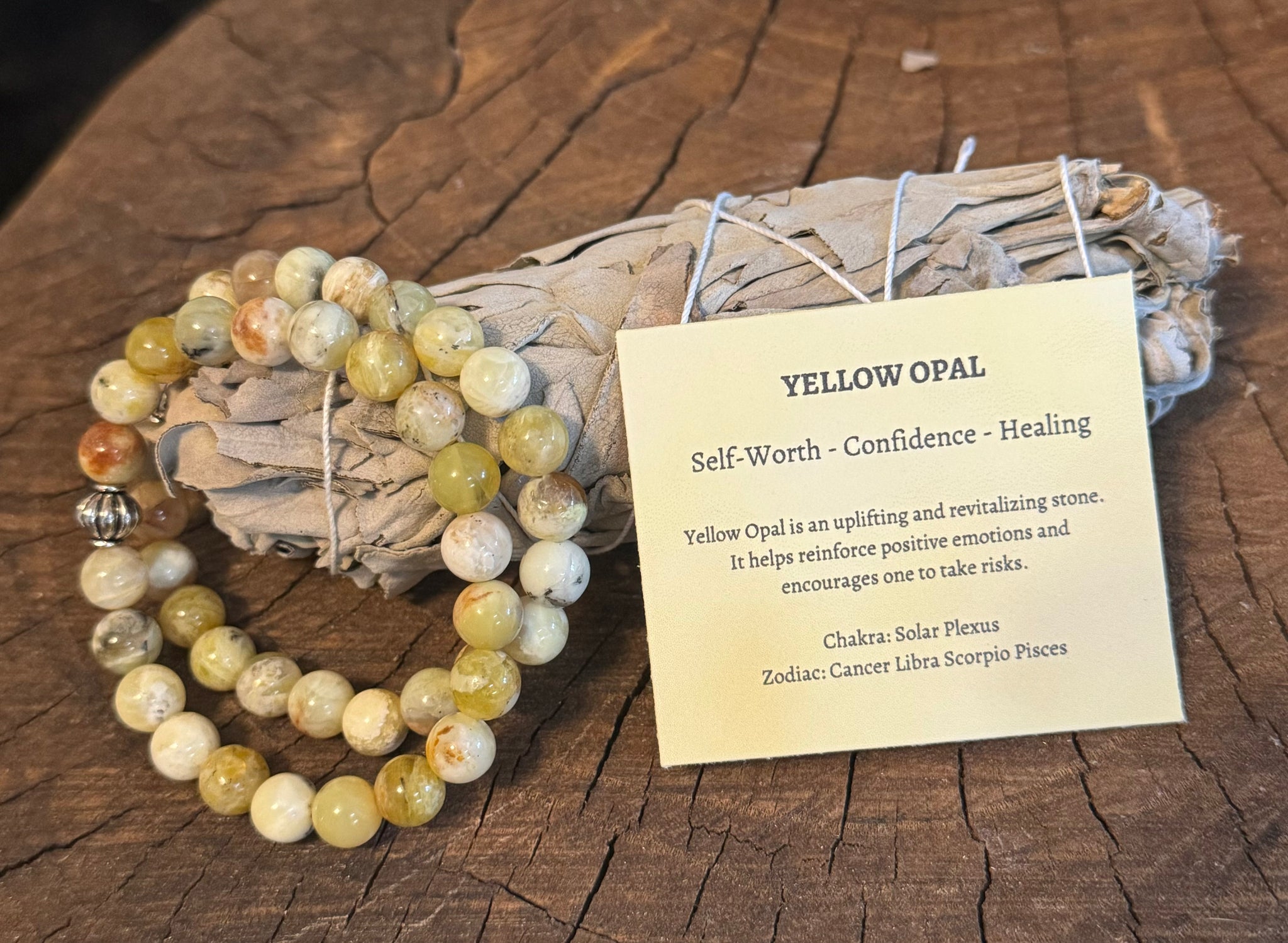 My Two Daughters Jewelry - A Yellow Opal Bracelet displayed alongside a card detailing its properties, placed on a wooden surface with a white sage bundle in the background.