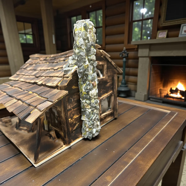 LB - Model cabin with stone chimney on a wooden surface in front of a fireplace.