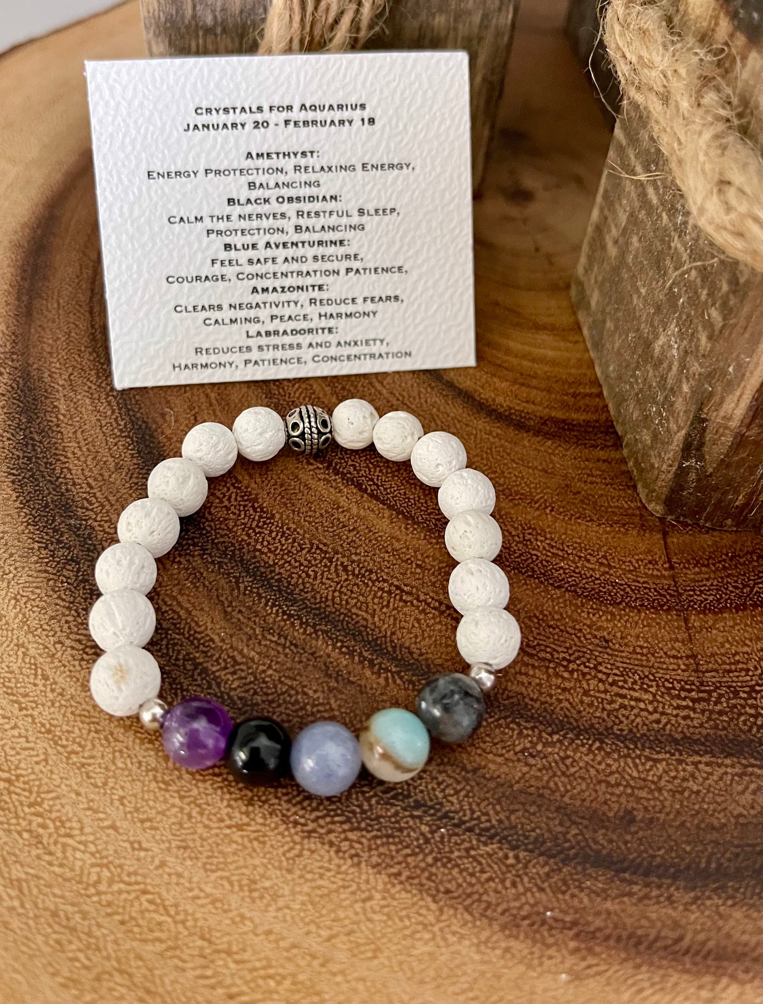 My Two Daughters Jewelry - An Aquarius Zodiac Bracelet with white and multicolored beads, displayed on a wooden surface with a card detailing the bracelet's properties.