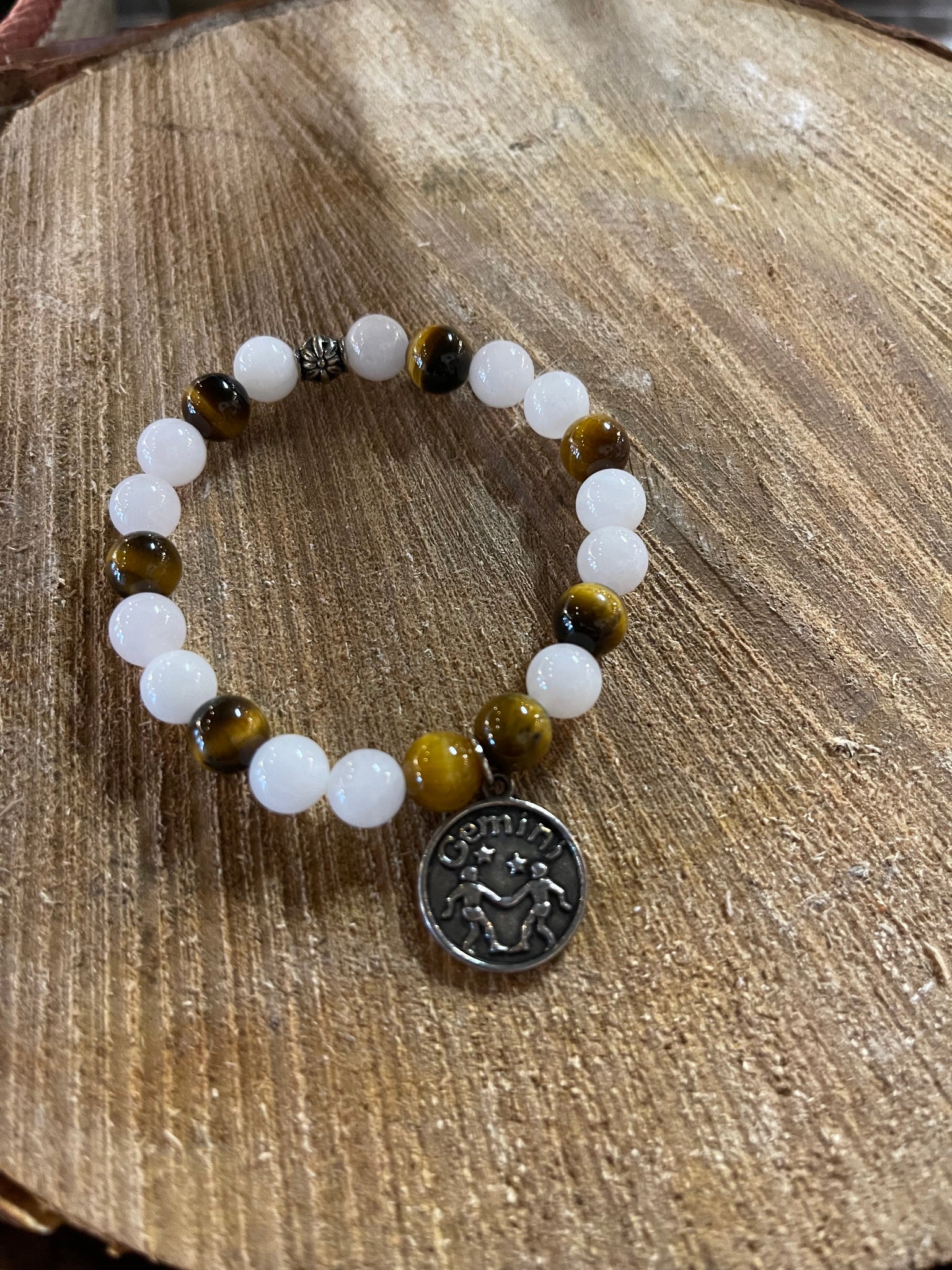 My Two Daughters Jewelry - A bracelet with white and yellow beads, and a silver-colored Gemini zodiac charm, displayed on a wooden surface.