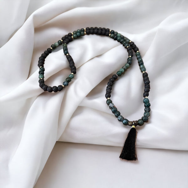 My Two Daughters Jewelry - A necklace made of black lava and green moss agate beads with a tassel, laid out on a white fabric surface.