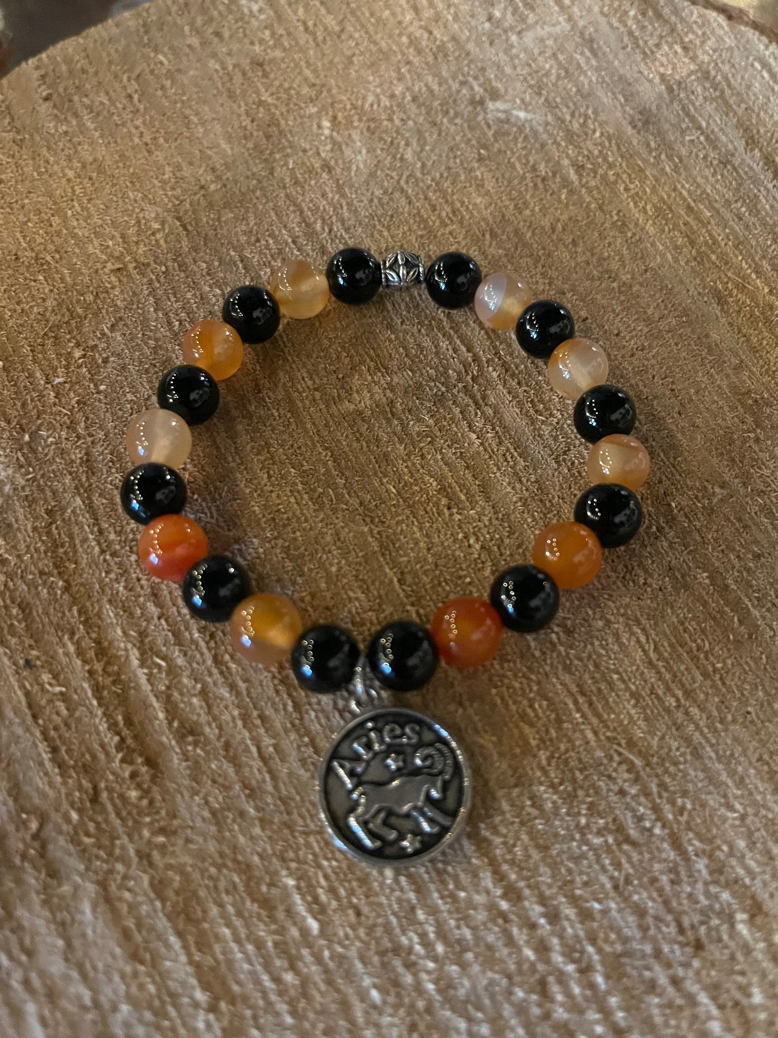 My Two Daughters Jewelry - A bracelet with black and orange beads, and a silver Aries zodiac charm, displayed on a wooden surface.